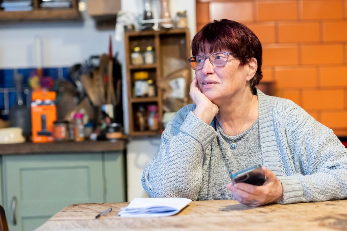 Frau über 60 sitzt nachdenklich mit dem Smartphone am Küchentisch